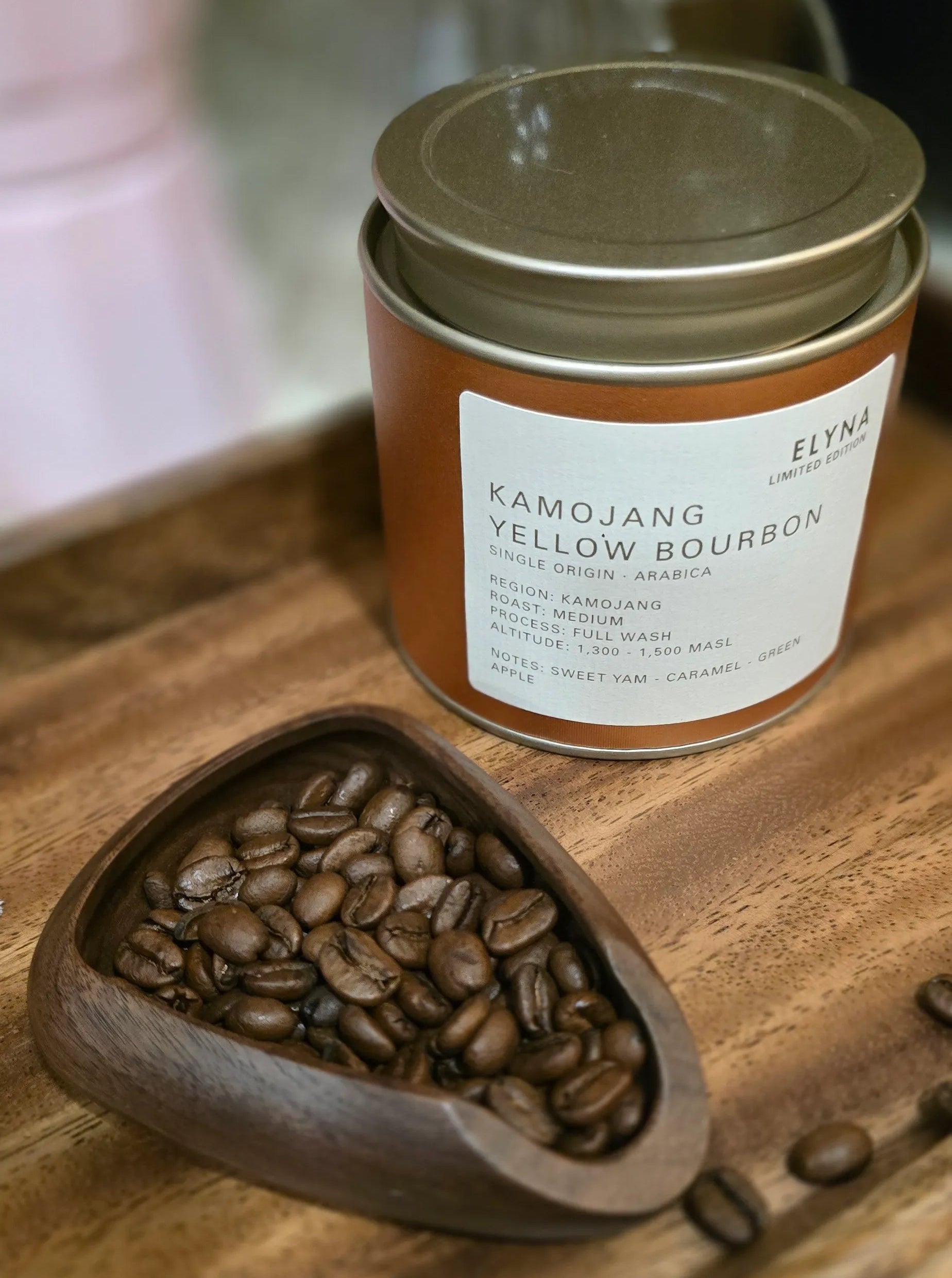 A jar of Elyna Kamojang Yellow Bourbon coffee beans sits on a wooden tray next to a small wooden scoop filled with roasted coffee beans.